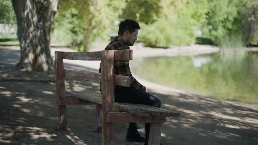 A man wearing a flannel shirt and dark pants sits alone on a wooden park bench facing a lake
