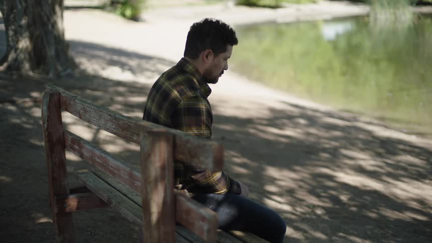 A man in a plaid shirt sits alone on a bench by a pond looking down sadly