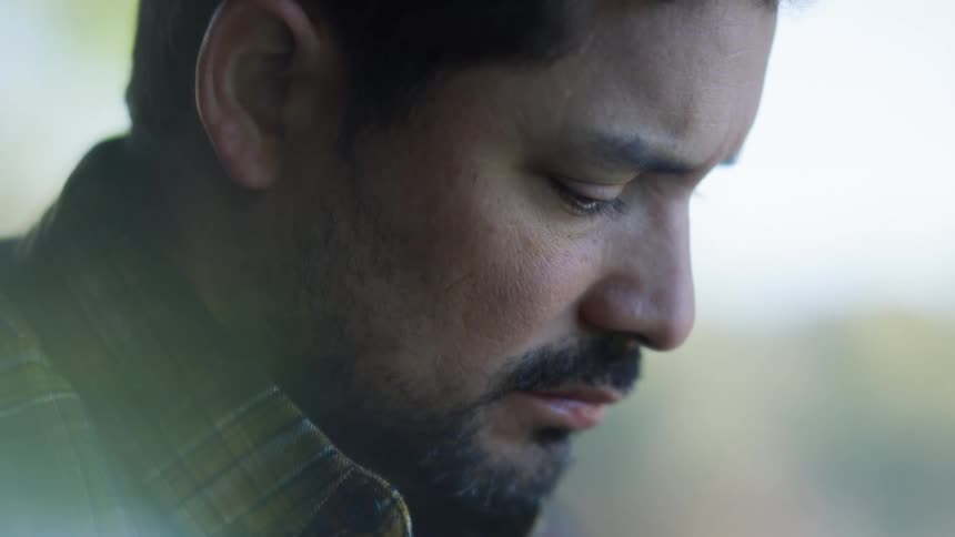 A man with a beard and a plaid shirt looks downward with a somber reflective expression the closeup shot captures his quiet moment of contemplation against a soft blurred background