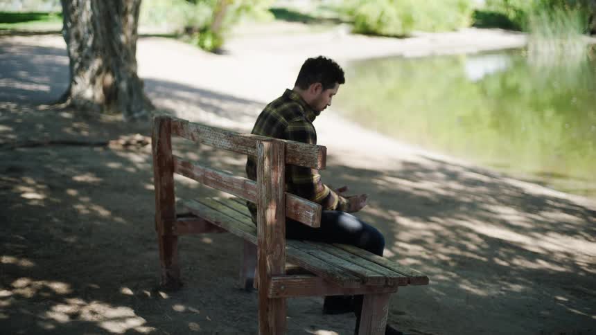Man sitting on a wooden bench looks at an object in his hands in an outdoor area beside a lake