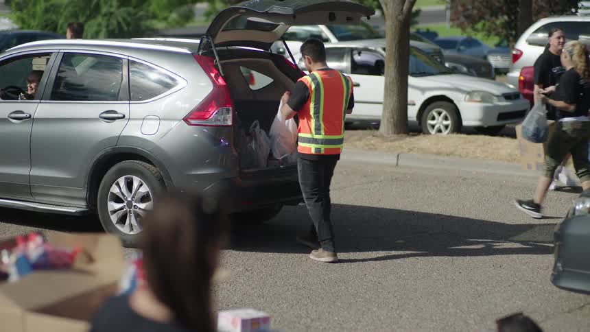 A wide shot shows a volunteer in a bright orange safety vest walking toward a gray car with its trunk open and placing two white plastic bags filled with groceries inside at a busy outdoor food distribution site