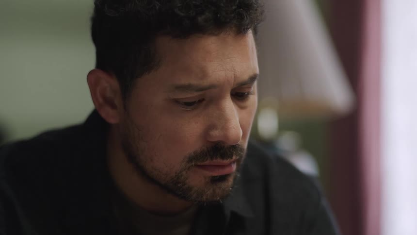Man seated indoors looks at something off frame with a focused expression
