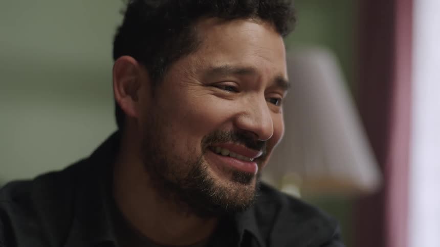 A young bearded latino man sits in a room at midday laughing in relief with tears of joy as he speaks to someone in bed