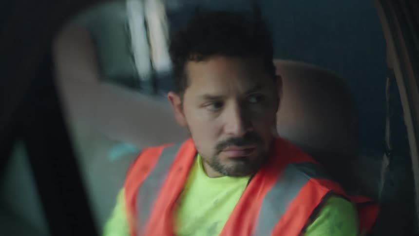 Young adult bearded man wearing a safety vest and a white sweater sitting on a vehicle in the rain with a sad facial expression then grabbing a helmet and making a motion to stand up indoors during night time