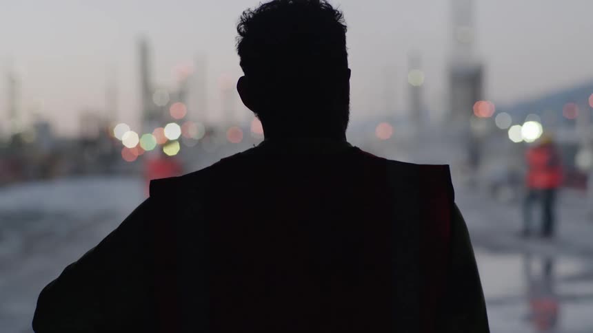 A silhouetted worker wearing a safety vest stands looking at an industrial site at dusk then he puts on his hard hat and begins walking toward the distant lights and structures of the facility