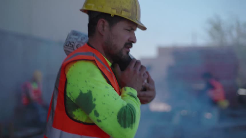 A construction worker wearing a safety vest and hardhat carries a cement sack on his shoulder while walking through a dusty outdoor job site during the day