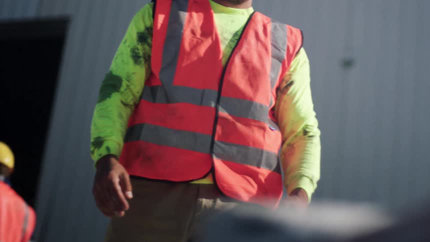 A latino cement worker in a hardhat carries a heavy bag of concrete on his shoulder