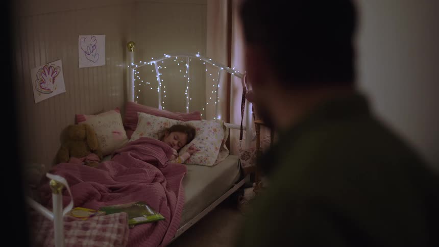 A man stands in a doorway and watches a young girl sleeping in a bed decorated with fairy lights pink blankets and drawings on the wall