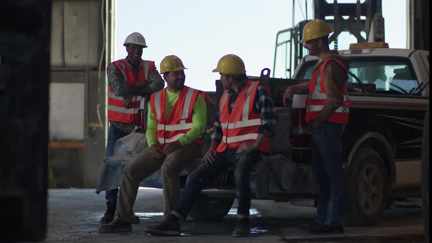 Four construction workers wearing safety gear sit on and stand around the tailgate of a white pickup truck inside a warehouse talking and laughing together during a work break