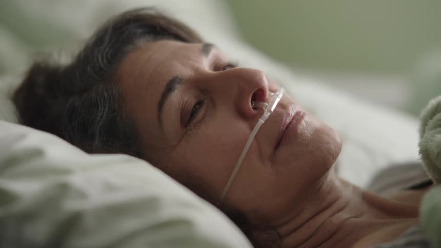 An older woman with a nasal cannula lies in a hospital bed and smiles