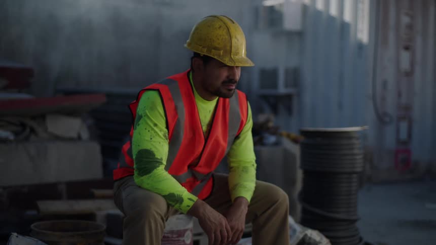 Exhausted latino construction worker sitting at a worksite wearing a hard hat and safety vest (frustrated)