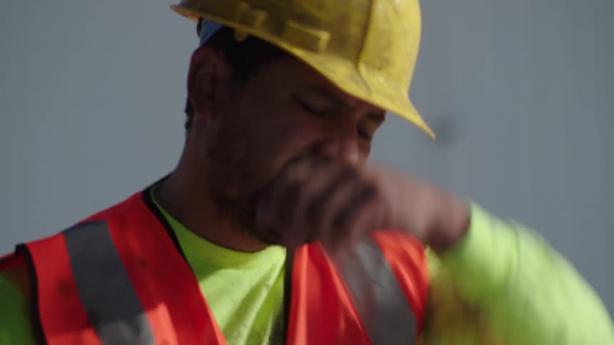 An exhausted latino construction worker carries a heavy cement bag on the shoulder at a work site