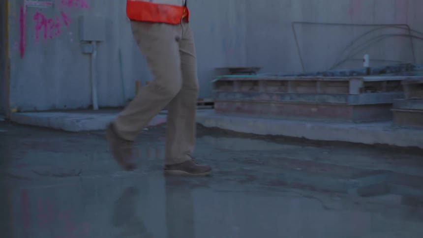A side view shows a construction worker in an orange safety vest and tan pants walking across a wet gray surface on a smoky construction site