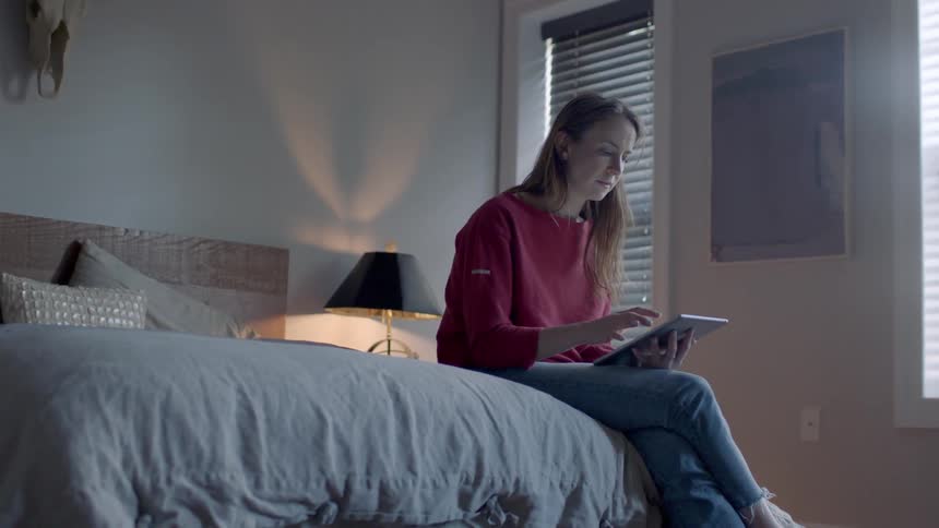 A woman sits on the bed browsing on a tablet