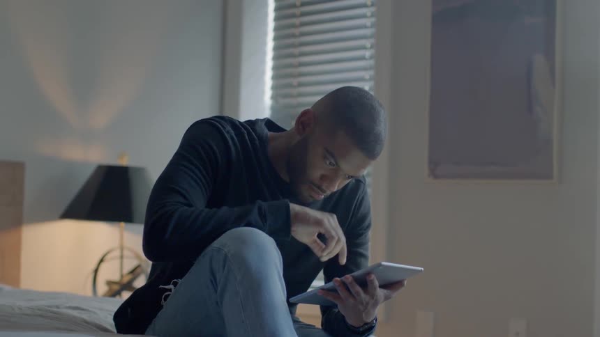 Man sitting on bed in bedroom is scrolling on an ipad tablet