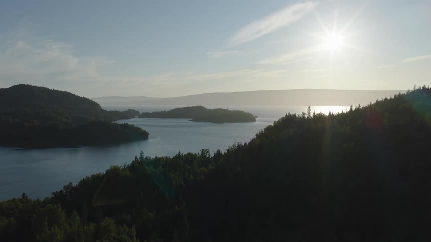 An aerial shot reveals a vast serene lake nestled among densely forested rolling hillsthe bright sun hangs low in the clear sky casting a shimmering golden reflection across the waters surface