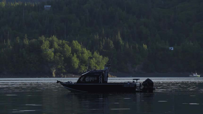 A black motor boat sits still on the calm surface of a lake at dusk while a dense evergreen forest covers the steep hills in the background and a second smaller boat passes slowly in the distance