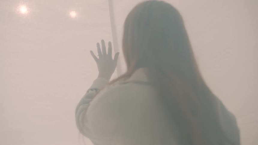 Young adult woman wearing white clothes touching a white curtain with her hand in a room with flickering lights indoors