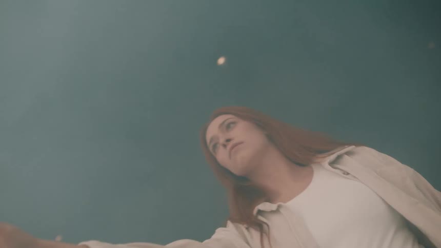 A woman with long hair stands in a smoky environment looking upward with a somber expression while reaching her hand toward the light as small embers float around her in a lowangle shot