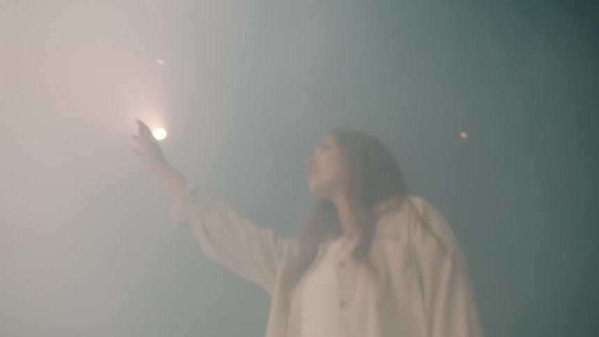 A woman stands in a dimly lit hazy room and reaches her hand upward toward a small bright light source on the ceiling while looking toward it