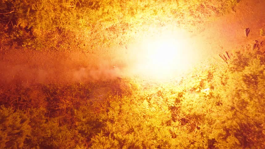 A topdown view at night shows a person riding a bicycle with a bright headlight down a narrow dirt path through a dense dark forest followed by a sudden intense orange flash that illuminates the surrounding trees and fills the air with smoke