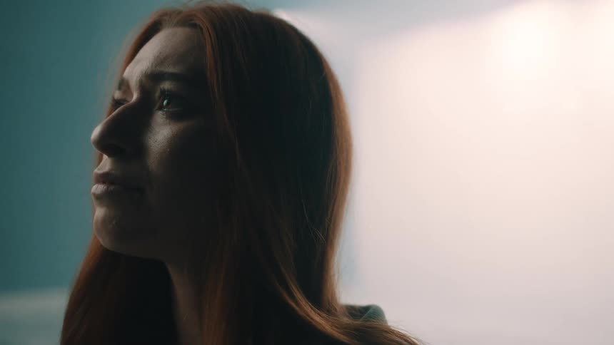 A woman with long red hair looks upward with a contemplative and emotional expression in a dimly lit studio while a soft white spotlight glows behind her against a blue background