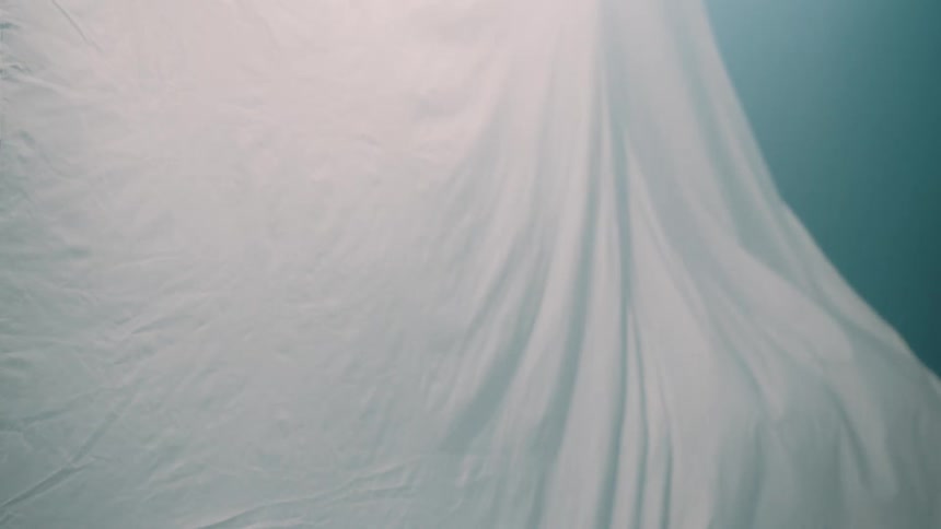 A white fabric sheet hangs and ripples gently against a light blue background illuminated from above