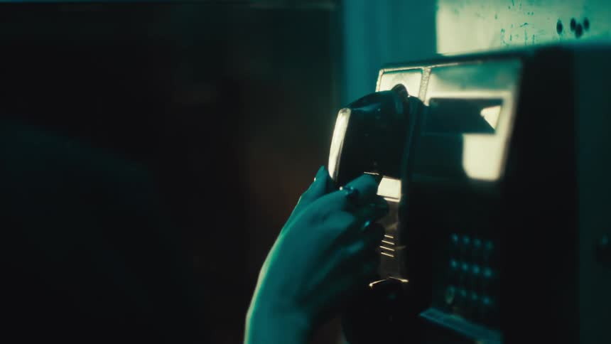 A person wearing rings reaches for a black payphone receiver in a dimly lit booth at night lifts the handset to their ear and prepares to make a call while the keypad glows in the background