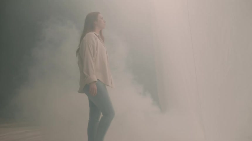 A young woman with long red hair walks through a room filling with white smoke her longsleeve shirt and jeans softening into the hazy atmosphere around her