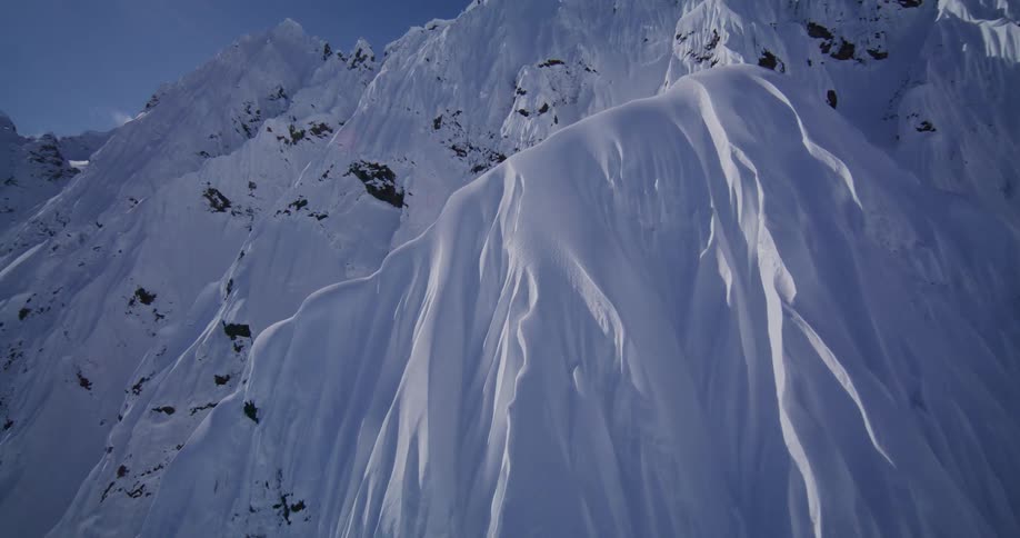 An aerial camera glides over a massive mountain range covered in thick white snow and ice capturing the sharp ridges and deep shadows of the jagged peaks under a clear blue sky and bright sunlight