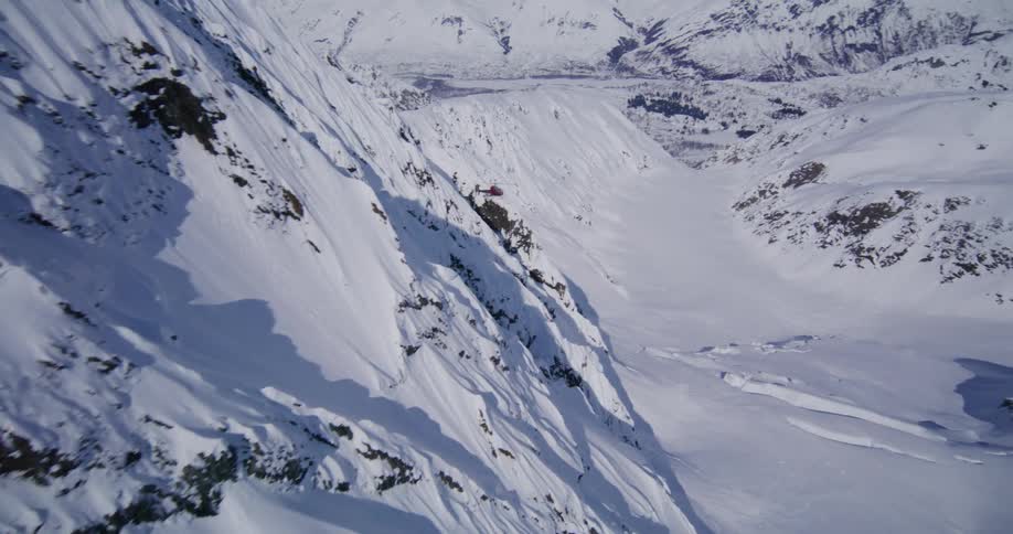 A red helicopter flies through a vast snowcovered mountain range and glides over a deep alpine valley