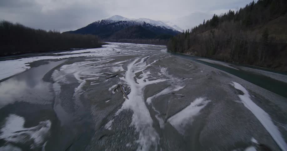 An aerial shot shows a vast braided river valley in winter with patches of snow and ice covering the gravel bars between turquoise channels of water flanked by dark forests and snowcapped mountains under an overcast sky