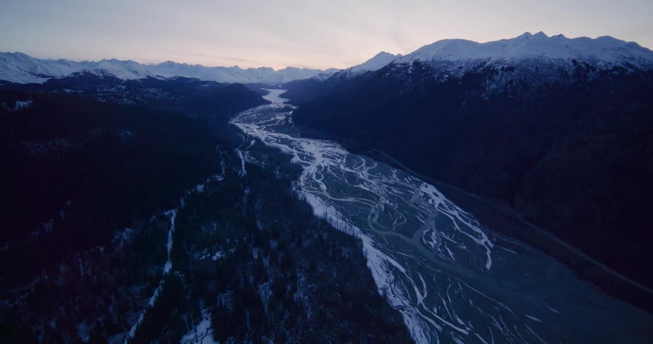 A stunning aerial view reveals a vast braided river winding through a deep mountain valley its icy tendrils stark against the landscape under a soft twilight sky