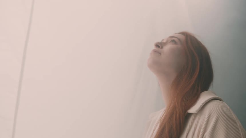 A red haired woman gazes upward while standing next to a white curtain