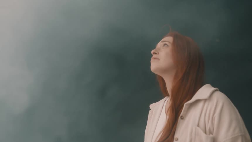 A young woman with long red hair stands in the room wearing a jacket looking up at the ceiling as white smoke fills the space and bright light reflects softly across her face
