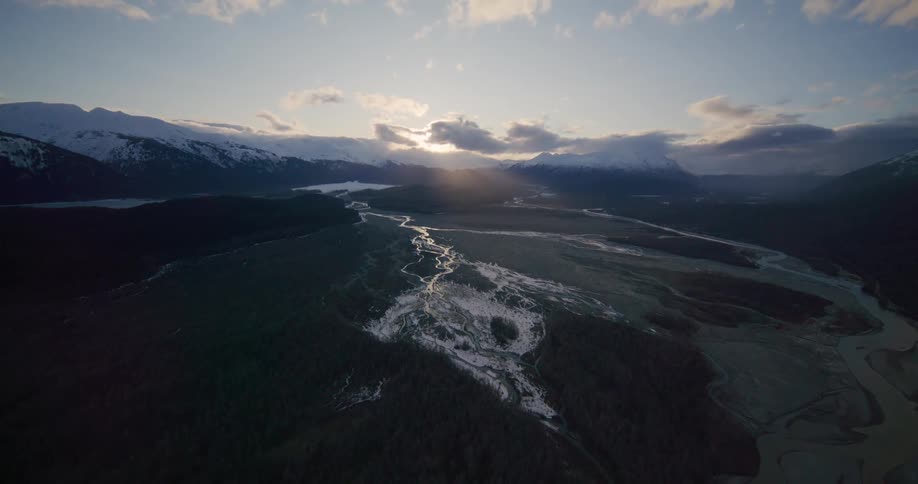 An aerial camera glides over a vast valley at sunset as the sun breaks through clouds above distant snowcapped mountains illuminating a winding river system that flows through the dark forested terrain and icy patches below