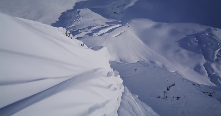 An aerial view plunges down the side of a steep snow covered mountain ridge