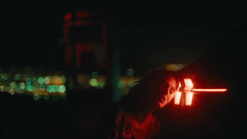 A view at night shows a woman leaning her head against a glowing red vehicle rear light with a red telephone booth and the blurred lights of a city in the background