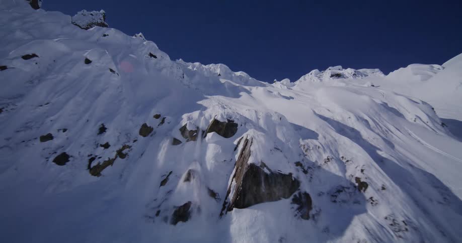 A steep snowcovered mountain ridge passing over jagged rock outcroppings and deep powder under a clear blue sky before pulling back to reveal the vast rugged alpine terrain