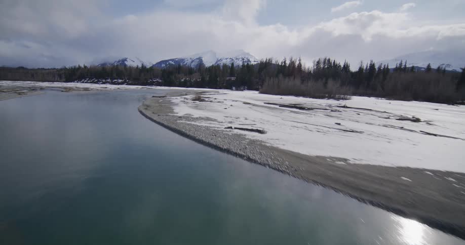 An aerial drone flies over a winding blue river flowing through a wide snowcovered valley toward distant snowcapped mountains under a bright cloudy sky