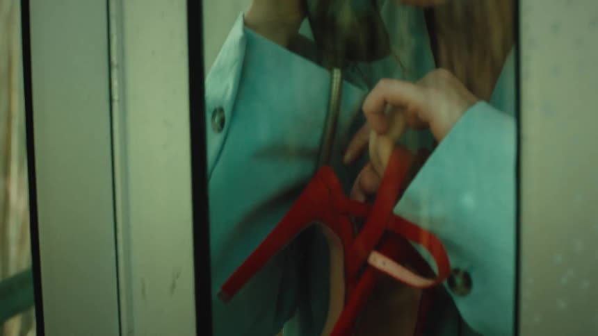 A woman in a lightblue coat talks on a handset inside a glass phone booth while holding a pair of red high heels her expression appearing distressed as she leans against the frame