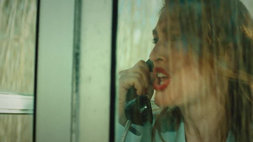 Woman making a phone call from a rural phone booth located near a field appears upset during the call