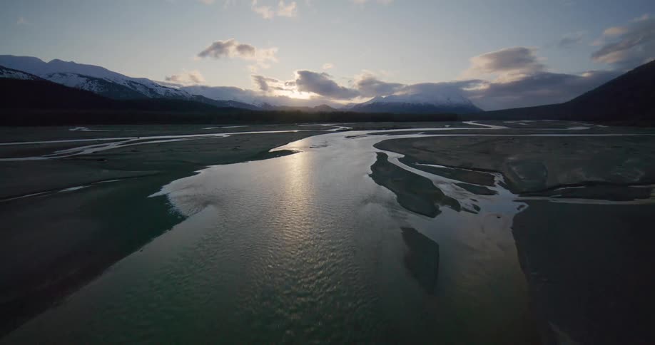 An aerial drone follows the curves of a silty braided river flowing through a wide glacial valley toward snowcapped mountains during a sunset
