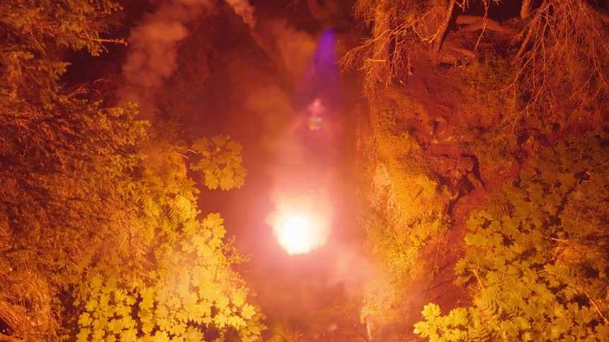 Fireworks burst beneath the forest at night briefly lighting up the surrounding trees and hills before fading back into darkness