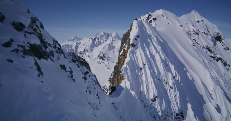 An aerial camera glides over sharp snowcovered mountain peaks and steep ridges under a clear blue sky revealing a vast winter wilderness with deep valleys and distant alpine ranges stretching toward the horizon