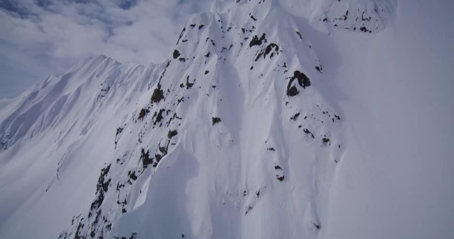 An aerial view sweeps rapidly past a rugged snow covered mountain ridge