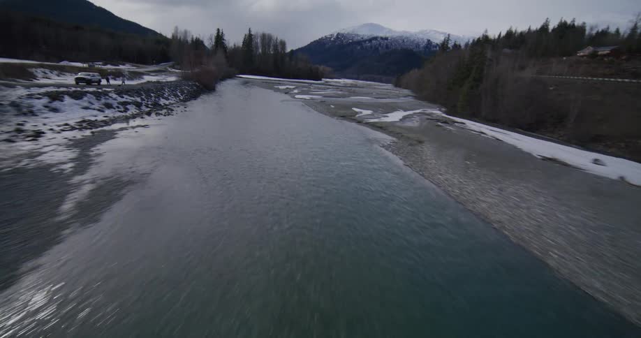 A drone flies over a concrete bridge and glides low across a cold river winding through a vast valley surrounded by snowdusted mountains and evergreen forests under an overcast sky