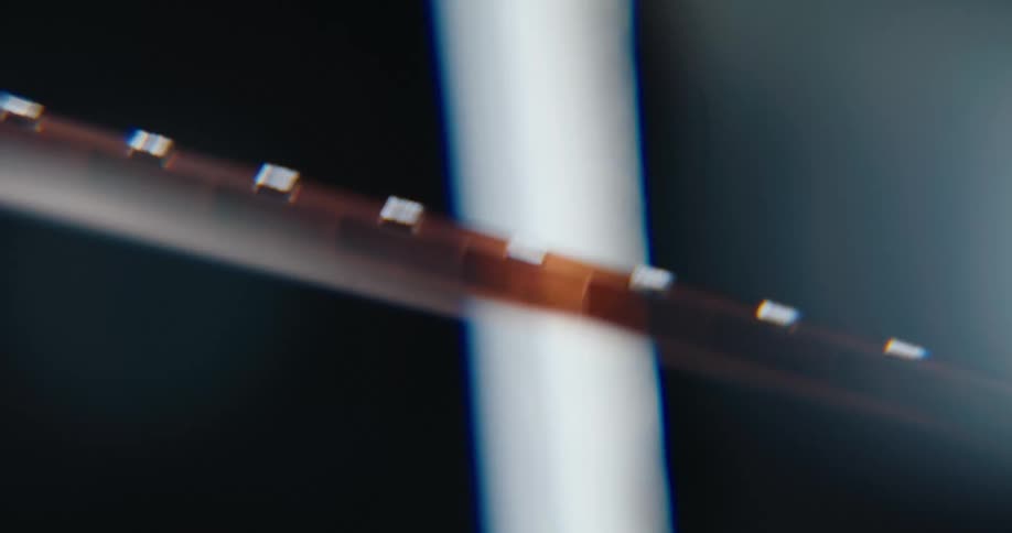 A macro closeup of a super 8mm film strip moving through a projector featuring visible sprocket holes and light flickering across the celluloid surface amidst a dark environment with subtle lens flares and dust