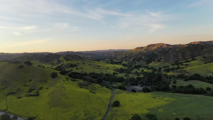 An aerial drone shot captures a vast landscape of vibrant green rolling hills and mountains during sunset featuring a winding road and a small utility structure nestled within the valley under a clear sky transitioning into evening light