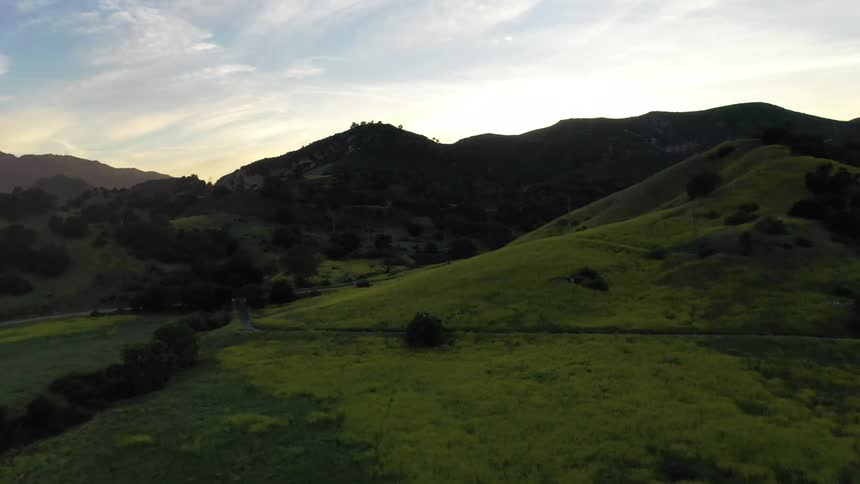 An aerial shot sweeps over rolling green hills and dark silhouetted mountains during a quiet dusk